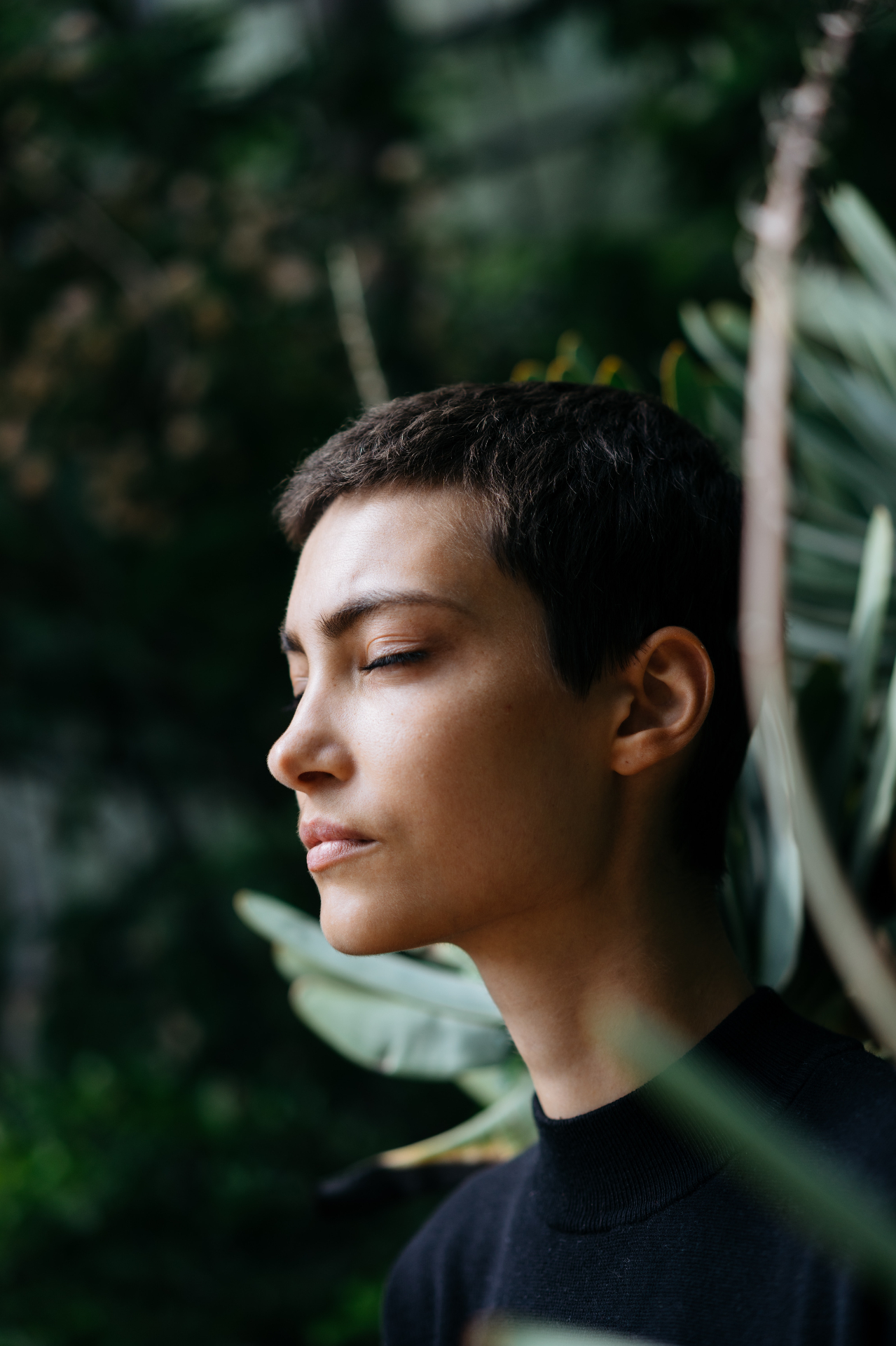 Woman with very short hair, eyes closed in profile against soft greenery